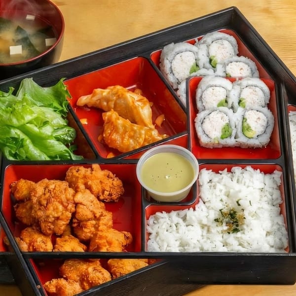 A bento box with sushi rolls, fried chicken, dumplings, rice, lettuce, and miso soup.