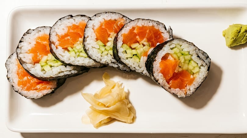Salmon and cucumber sushi rolls with pickled ginger and wasabi on a white rectangular plate.