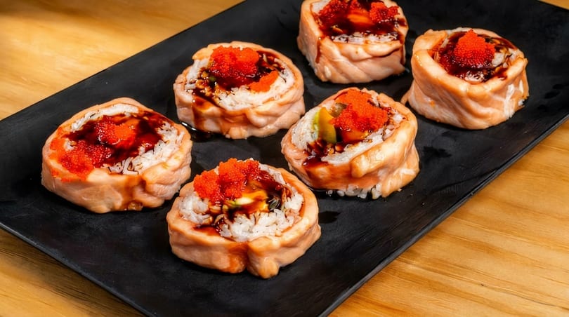 Six salmon sushi rolls with rice, roe, and dark sauce on a black rectangular plate.