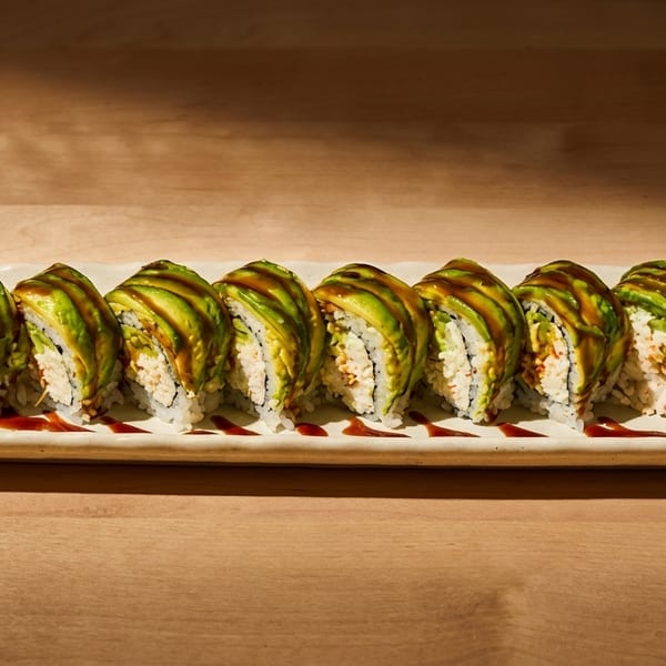 Eight avocado-topped sushi rolls with sauce, wasabi, and ginger on a white rectangular plate.