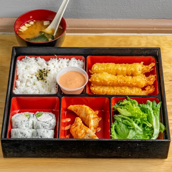 A bento box with rice, fried shrimp, sushi, dumplings, lettuce, and a side of miso soup.