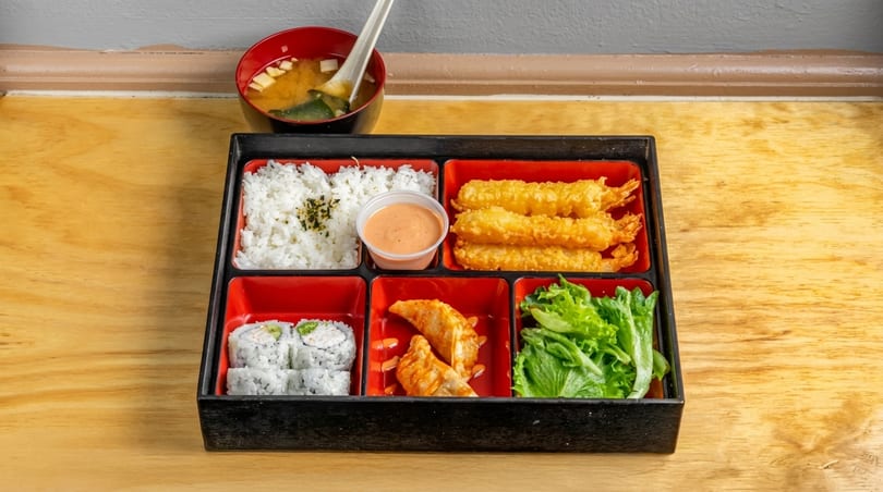 A bento box with rice, fried shrimp, sushi, dumplings, lettuce, and a side of miso soup.