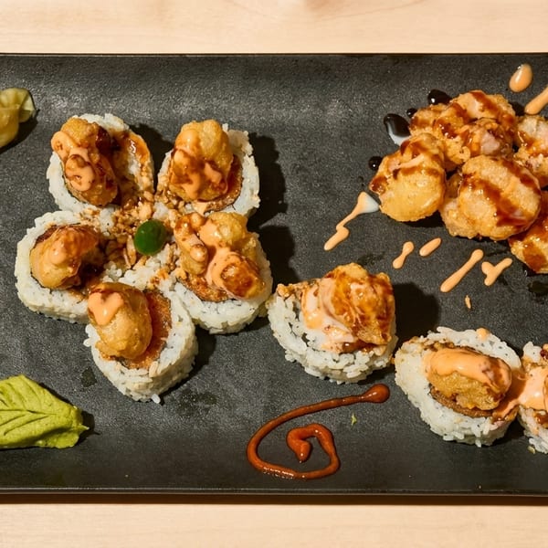 Sushi rolls with fried toppings and various sauces, alongside pickled ginger and wasabi on a black plate.