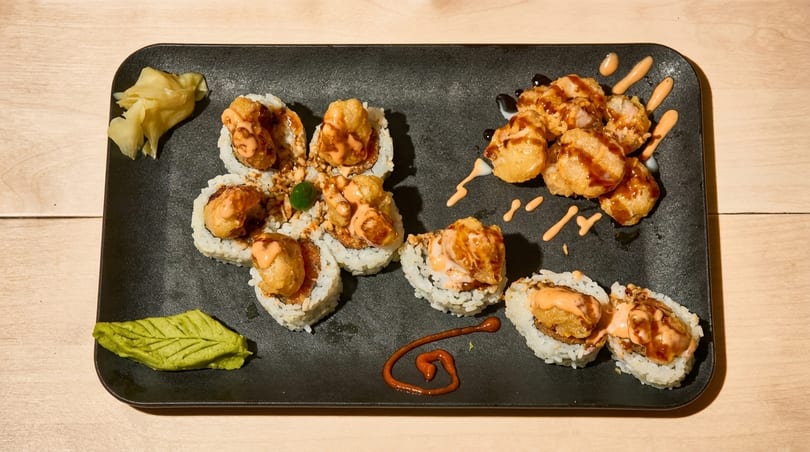 Sushi rolls with fried toppings and various sauces, alongside pickled ginger and wasabi on a black plate.