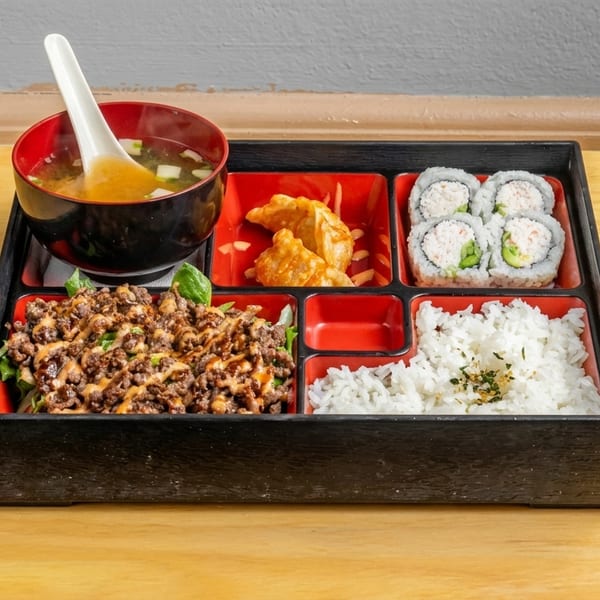 A bento box with miso soup, gyoza, California rolls, rice, and bulgogi over greens.