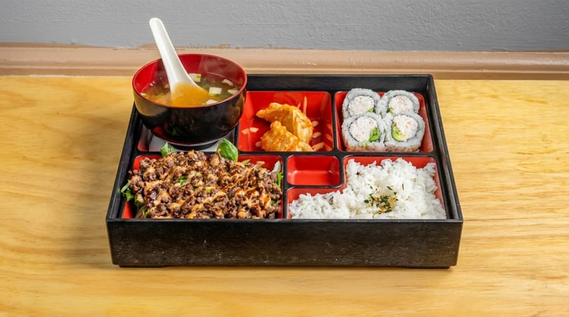 A bento box with miso soup, gyoza, California rolls, rice, and bulgogi over greens.