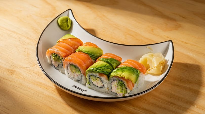 A crescent-shaped plate holds six sushi rolls topped with salmon and avocado, with wasabi and ginger.