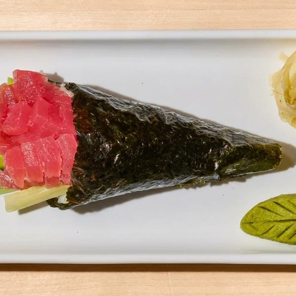 Tuna hand roll with cucumber, pickled ginger, and leaf-shaped wasabi on a white rectangular plate.