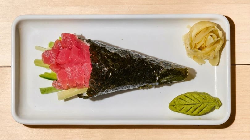 Tuna hand roll with cucumber, pickled ginger, and leaf-shaped wasabi on a white rectangular plate.