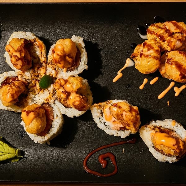 Sushi rolls and fried shrimp tempura with sauces, pickled ginger, and wasabi on a black plate.