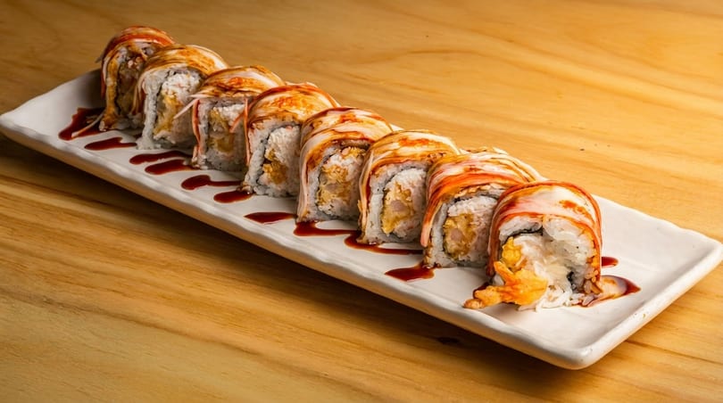 A white rectangular plate holds a row of ten sushi rolls drizzled with dark sauce on a wooden table.