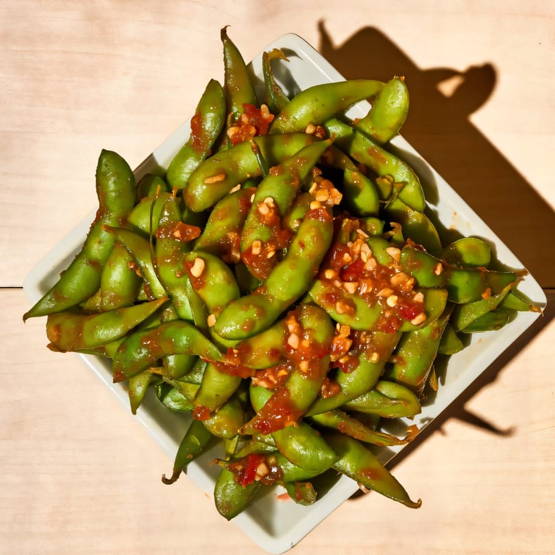 A white square bowl filled with green edamame pods topped with a spicy red sauce and garlic bits.