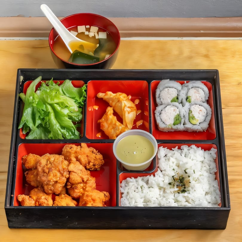 Sushi Go bento box with sushi rolls, fried chicken, dumplings, rice, salad, and miso soup.